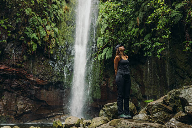 Front view of handsome female traveller in black sport clothes hiking in the tropical forest contemplating scenic waterfall on Madeira island