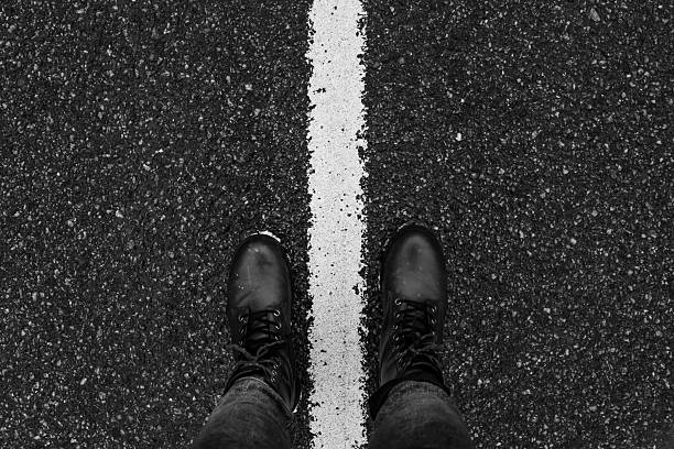 A black and white image of a person wearing boots standing on a dividing road line, symbolizing direction, choice, and perspective with a textured asphalt background