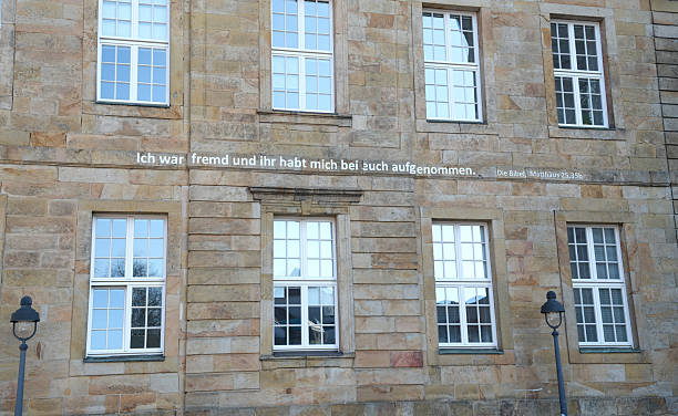 Stone building facade featuring symmetrical white-framed windows with multiple panes. A sentence in from the bible in German is inscribed across the wall: I was a stranger and you invited me in. The building is from the 18th century and build in the style of Neoclassicism