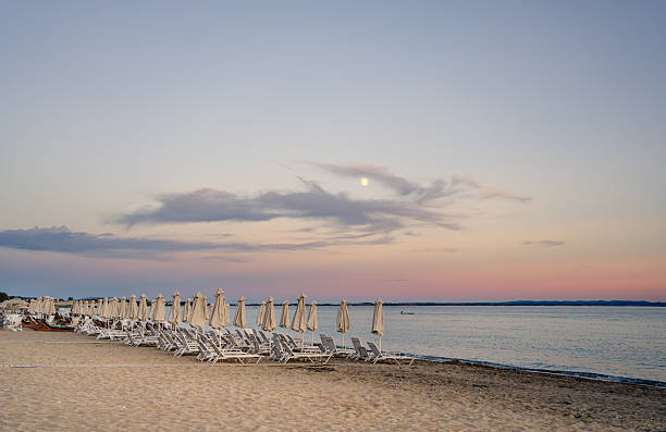 White sunbeds and umbrellas lining a sandy beach, basking in the warm hues of sunset, with the moon and clouds softly illuminating the sky
