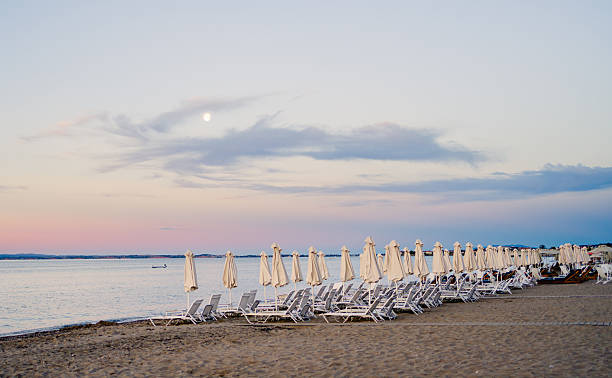 White sunbeds and umbrellas lining a sandy beach at sunset, with the moon rising in a pastel sky filled with soft pink and blue hues