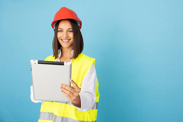 istock Young female engineer wearing safety helmet and vest using digital tablet, isolated on blue background 2212607213