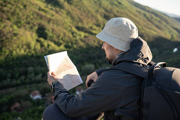 A traveler studies a map while enjoying the breathtaking mountain scenery during sunrise, wearing gear suitable for outdoor exploration. The peaceful environment captures a sense of adventure, calmness, and nature's beauty.