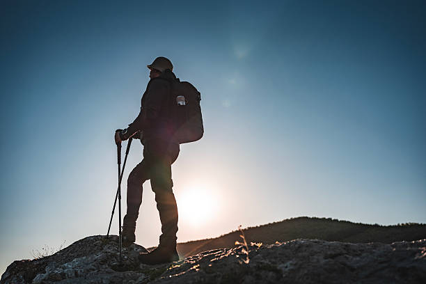 A hiker silhouetted against the early morning sunlight, standing atop a rocky hill with trekking poles and a backpack, embracing adventure and outdoor exploration in nature.