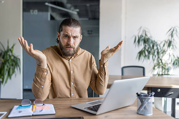 istock Confused man in office using laptop and raising arms in question 2212617545