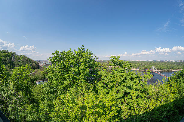 istock Landscape with lush green trees, a bridge over water connecting two areas, distant cityscape, clear blue sky, wide angle view. 2212625457