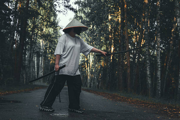istock A martial artist in a conical Asian hat practices fighting techniques with a katana sword on a road in the forest. Katana training in the forest against the backdrop of autumn trees. 2212630972