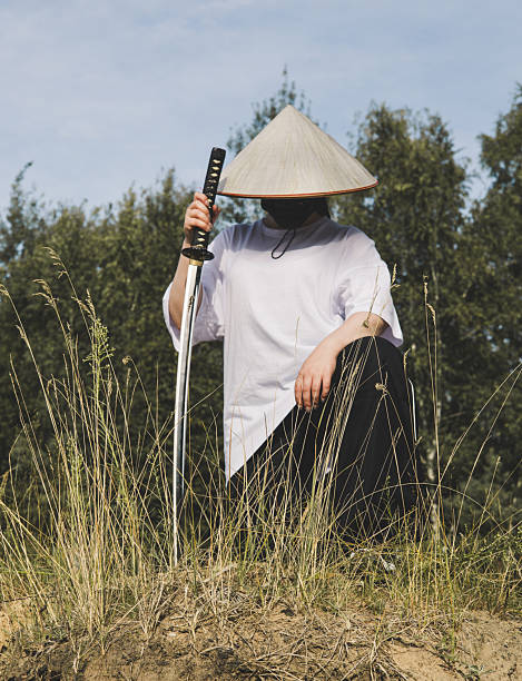 istock A warrior katana on the river bank. A man dressed as a samurai, with a conical hat and mask, kneels in nature holding a katana, creating a striking contrast between tradition and modernity. 2212632070