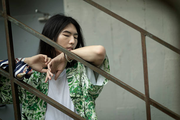Fashionable young man with long hair leaning against a railing, gazing thoughtfully away while exuding a calm and stylish vibe in an urban setting