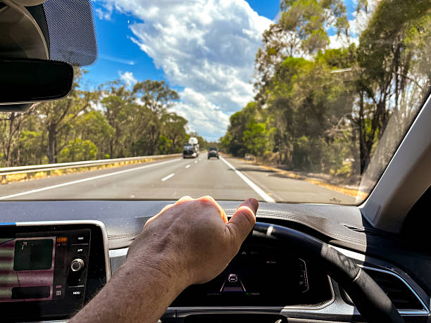 istock A wide-angle view through a car’s windscreen reveals a highway through a forested area, framed by the interior, steering wheel, and driver’s hands under a clear blue sky with fluffy clouds 2212649539
