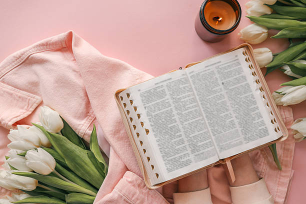 istock Open Bible held in hands over a pink background, surrounded by white tulips, a lit candle, and a pink denim jacket, creating a peaceful devotional setting 2212653697