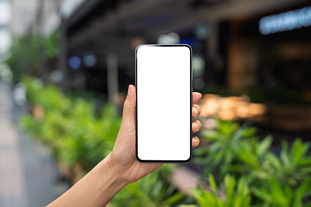 A female hand holds a smartphone with a blank white screen in focus, against a blurred background of an outdoor café. Perfect for mobile app or advertisement mockups.