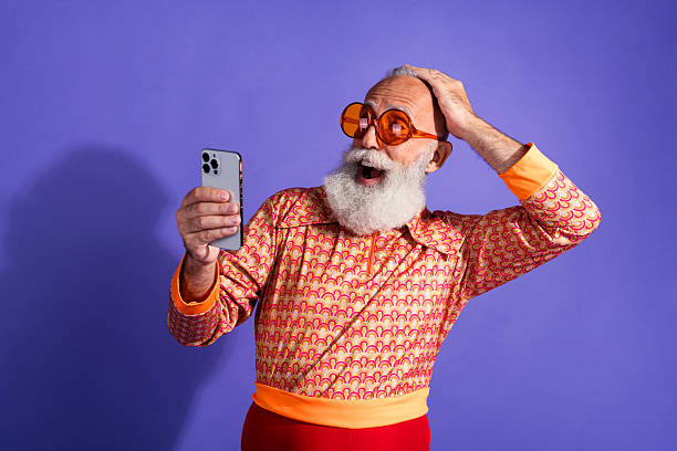 istock Elderly stylish man with beard and retro outfit ecstatic while looking at smartphone against fun vibrant purple backdrop 2212691440