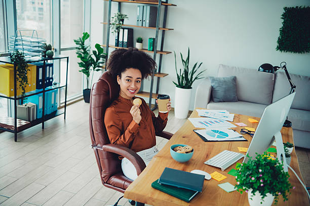 istock Young professional woman enjoying coffee and snacks in a bright modern office setting with plants and organized workspace. 2212693303