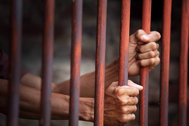 istock Hands Gripping Metal Bars in Dark Setting of a Prison or Cell 2212693478