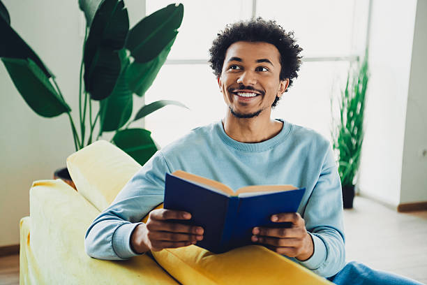 istock Smiling young man enjoys reading a book while relaxing on a couch in a modern apartment with stylish interior decor. 2212694503
