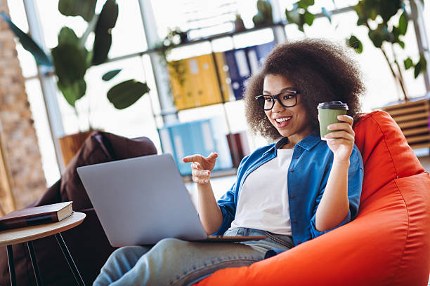 istock Young professional woman using a laptop while sitting on a bean bag, enjoying coffee, in a modern and stylish office space 2212695168
