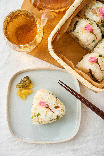 Homemade Japanese rice balls (onigiri) with spring vegetables, delicately topped with salted sakura blossoms, served in a bamboo basket on a bright spring day.