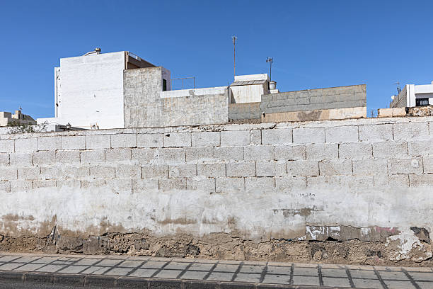 A weathered stone wall stands in Arrecife, Lanzarote, showcasing a mix of concrete blocks and natural stones in varying shades of gray and brown. Behind the wall, several buildings with flat roofs and rooftop structures, including antennas, add to the urban landscape. The clear blue sky contrasts with the muted tones of the architecture, highlighting the blend of traditional and modern elements in this coastal city. The image captures a quiet yet characteristic scene of Arrecife’s built environment.