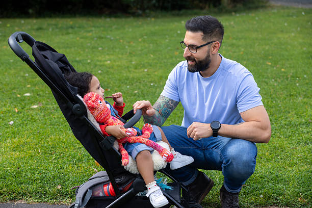 A medium shot of a father kneeling next to his baby in a pram at the park talking to her. He looks happy and caring, while the baby sits calmly in the stroller. The background is filled with greenery and a peaceful path, creating a warm, everyday moment of family life.\n\nVideos are available similar to this scenario.