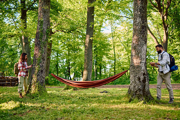 Young couple of hikers preparing an orange hammock, securely tying it between two trees in a lush forest on a bright summer day, enjoying the tranquility of nature and each other's company