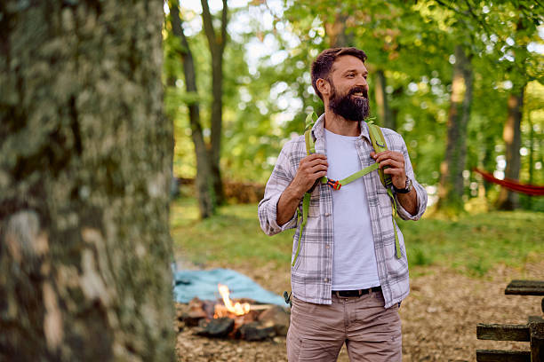 Happy camper wearing a backpack stands near a crackling bonfire in a serene forest, soaking in the tranquility and joy of nature during a memorable camping trip