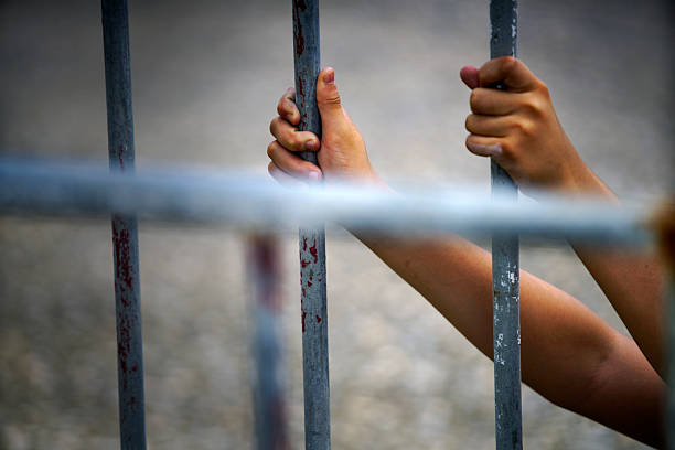 istock Hands Gripping Metal Bars Behind Prison Fence in Dim Light 2212731952