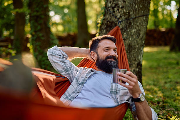 Bearded man relaxing in hammock drinking coffee in forest