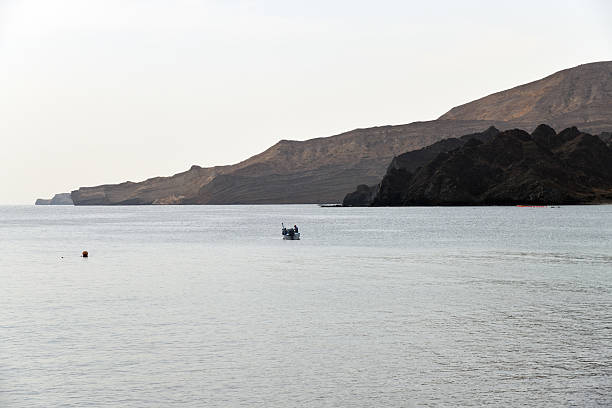 The coastline of Oman features sparkling blue waters contrasting with the rocky cliffs and majestic mountains creating a serene and picturesque landscape under bright sunlight.