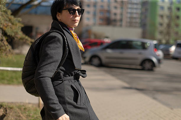 A man who is wearing dark sunglasses and a stylish black overcoat is taking leisurely steps as he walks down the sidewalk