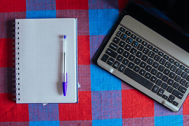 A notebook and pen are on a checkered table next to a laptop. The notebook is open to a page with a pen on it. The laptop is turned off and placed on the table