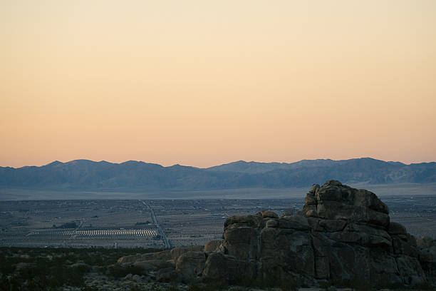 In the fading light of dusk, scattered buildings of a desert town appear in the distance, with solar panel installations reflecting the last rays of sun. The scene highlights the balance between human development and the vast openness of arid landscapes, evoking themes of sustainability, desert living, and renewable energy.