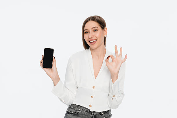 istock Smiling Woman Showing Smartphone and Making OK Gesture on White Background 2212772576