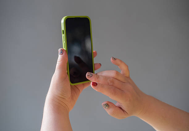 Close-up of hands using a smartphone with a bright green protective case against a neutral gray background. The person has painted fingernails with some decorative nail art. One hand holds the device while the other interacts with the screen. The smartphone display appears dark. The image captures the everyday gesture of mobile device operation with selective focus on the hands and phone.