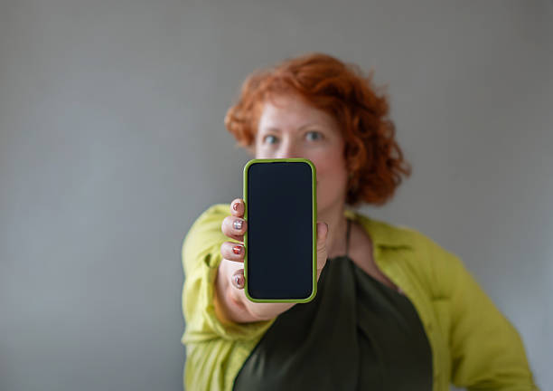 48-year-old woman with curly red hair holds up a smartphone with bright yellow case toward the camera. The subject wears a lime green cardigan over dark clothing and stands against a plain gray wall. The phone screen is turned toward the viewer in a close-up shot with selective focus on the smartphone and hand.