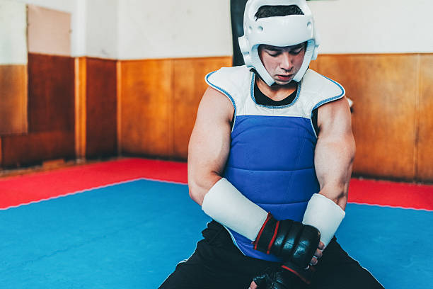 Male martial artist sits on a red and blue mat in a gym, adjusting his protective arm and shin guards before a training session. He is dressed in a blue and white padded vest and black shorts, demonstrating focus and readiness for combat sports practice.