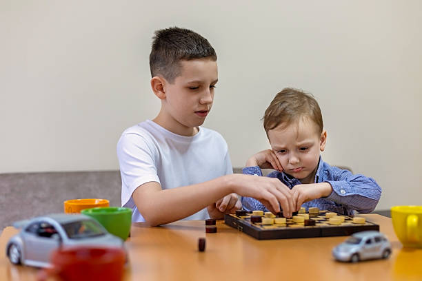 Two boys playing checkers at a table, with the younger one looking disappointed while the older one makes a move