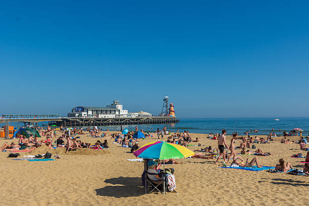 West Cliff Beach, Bournemouth, UK - April 30th 2025: Someone sitting under a parasol on the beach surrounded by people sunbathing in front of Bournemouth Pier.