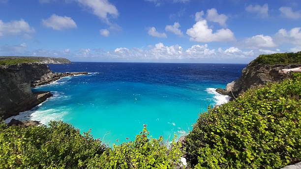Rugged coastline with dramatic limestone cliffs and an arch formation overlooking the deep blue sea. Waves crash against the rocks, creating white foam. Sparse greenery grows atop the cliffs, contrasting with the bright blue sky dotted with fluffy clouds. The natural archway and rocky terrain highlight the region's geological features.