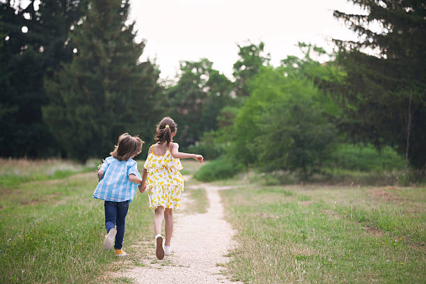 Two little girls wearing summer dresses walk arm in arm along a sunny forest trail
