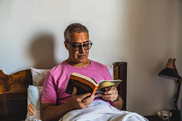 istock Middle aged white man is sitting on his bed reading a book with soft sunlight illuminating. 2212802336