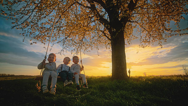 Three children enjoying an outdoor activity on a tire swing under a tree in summer. The image conveys a sense of fun, friendship, and the joys of spending time in nature during childhood.