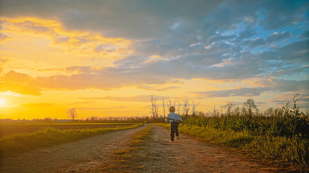Young Child Running Joyfully Along a Dirt Road in the Countryside at Sunset,Carrying a Stick and Basking in the Warm,Golden Light of the Evening Sky