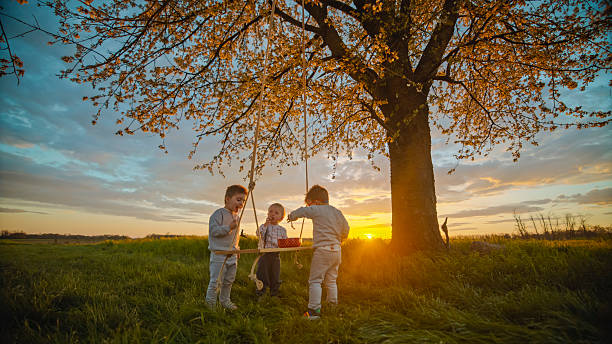 Three Young Children Playing on a Wooden Swing Beneath a Blossoming Cherry Tree at Sunset,Surrounded by a Serene Landscape That Embodies the Joy of Childhood and Nature's Beauty