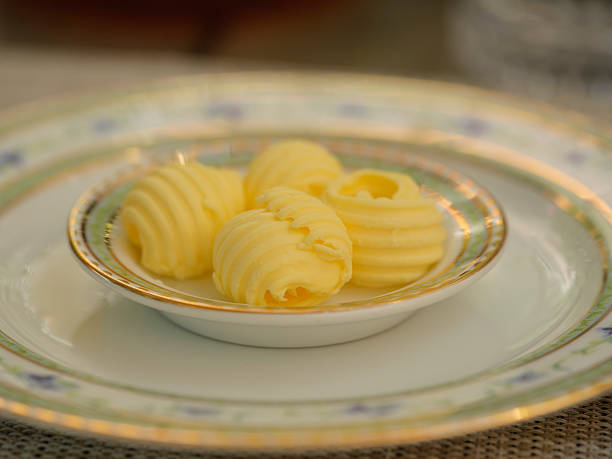 Four delicately textured butter curls are elegantly placed on a fine plate with an intricate floral design, capturing an ambiance of culinary precision and refined dining. A soft focus enhances the inviting aesthetic.
