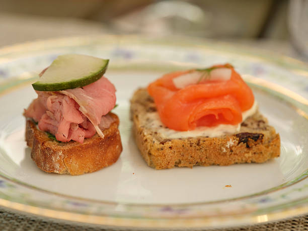 Two gourmet appetizers presented on a decorative plate. One features roast beef with cucumber on crusty bread, and the other showcases smoked salmon atop herbed cream cheese. Ideal for culinary and dining visuals.