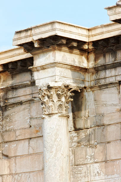 A close-up of a classical architectural stone column featuring an ornate Corinthian capital, situated against a weathered stone wall. The composition highlights its historical artistry under gentle sunlight.