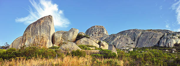 This scenic composition showcases impressive granite boulders nestled amidst verdant vegetation, highlighted under a vibrant blue sky adorned with wispy clouds. A tranquil, nature-embracing ambiance emanates from this scenic landscape.