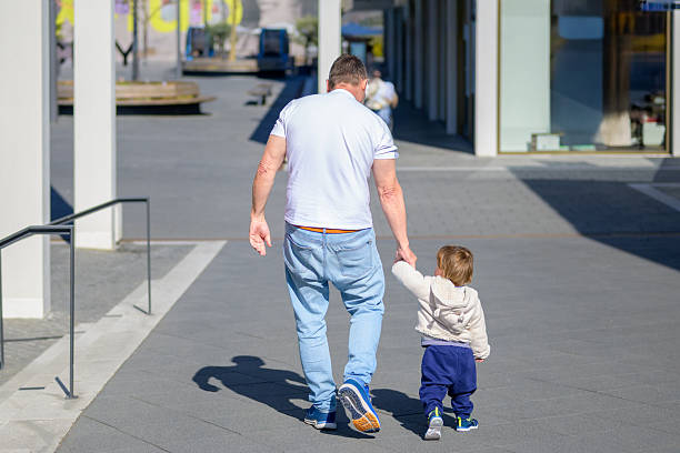 A grandfather and his young grandchild walk hand in hand through a modern urban plaza on a sunny day.