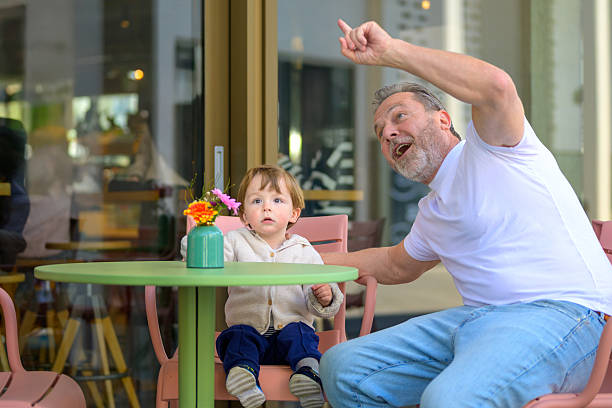A cheerful grandfather points excitedly toward the sky while sitting at an outdoor café with his young grandchild.
